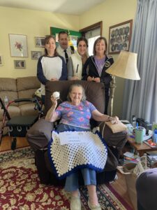 Alice is seated on a brown recliner. She holds a flower in her hand and has a knitted blanket on her lap. Alice's family is lined up behind her.