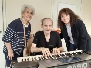 Marge, white woman with grey hair standing, next to John, white man with grey hair, who is seated hands on the piano. Next to them on the right is John's sister Pat who is standing and leaning in for the picture.